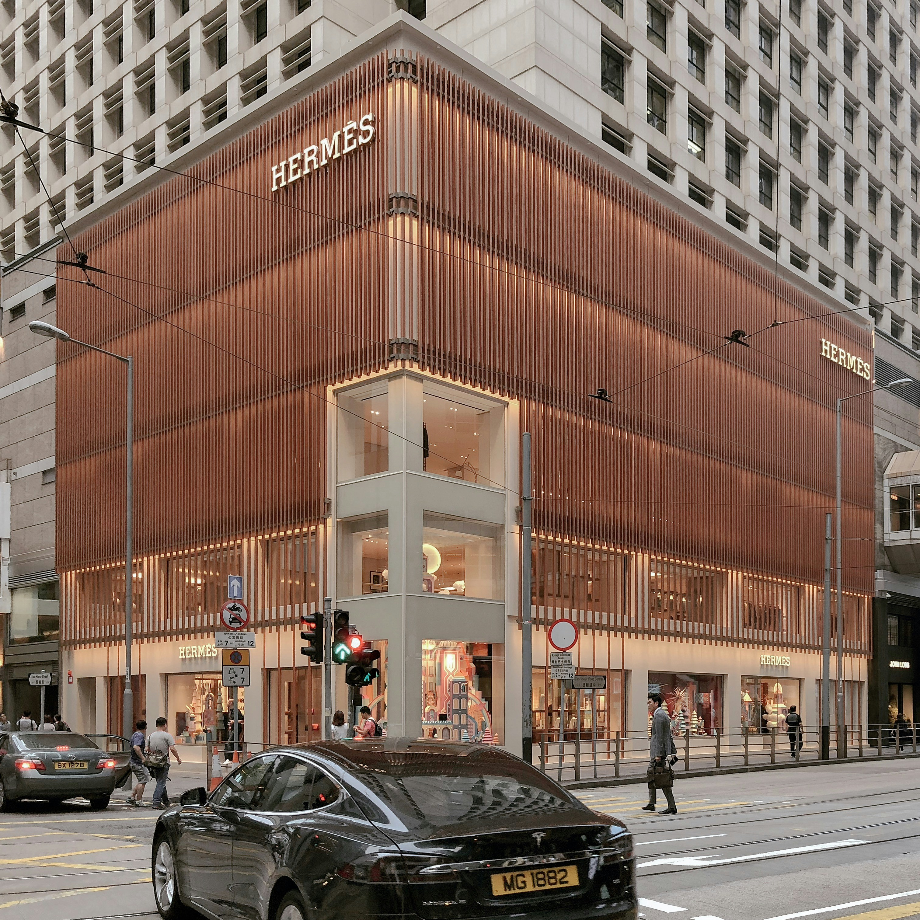 Corner view of Hermès store with vertical slat facade and a black Tesla car in front on city street.